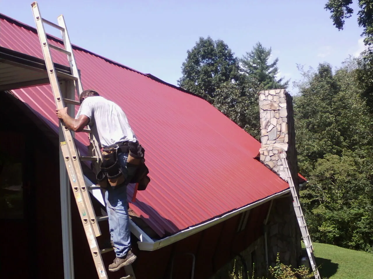 Roofer on a ladder beside a red metal roof and stone chimney — Soffit & Fascia Repair by Roofing Brothers in Yacolt